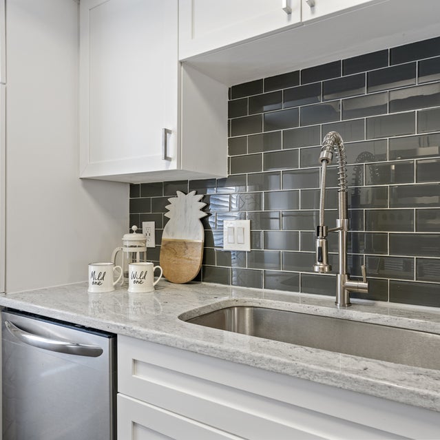 Andover At Crabtree Apartments - Kitchen With Farmhouse Sink, White Cabinetry, And Dark Gray Subway Tile Backsplash